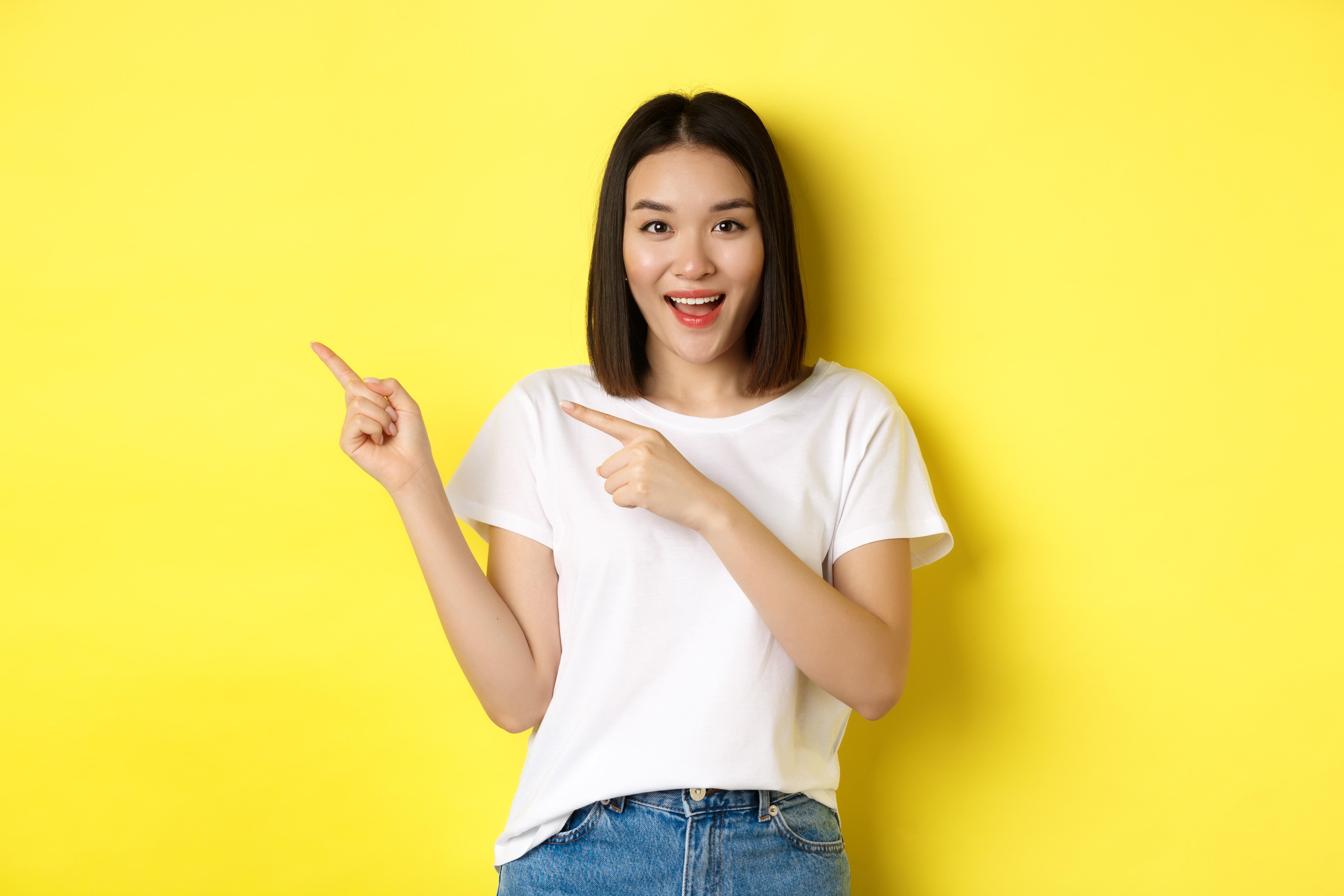 Beauty and fashion concept. Beautiful asian woman in white t-shirt pointing fingers left, standing over yellow background