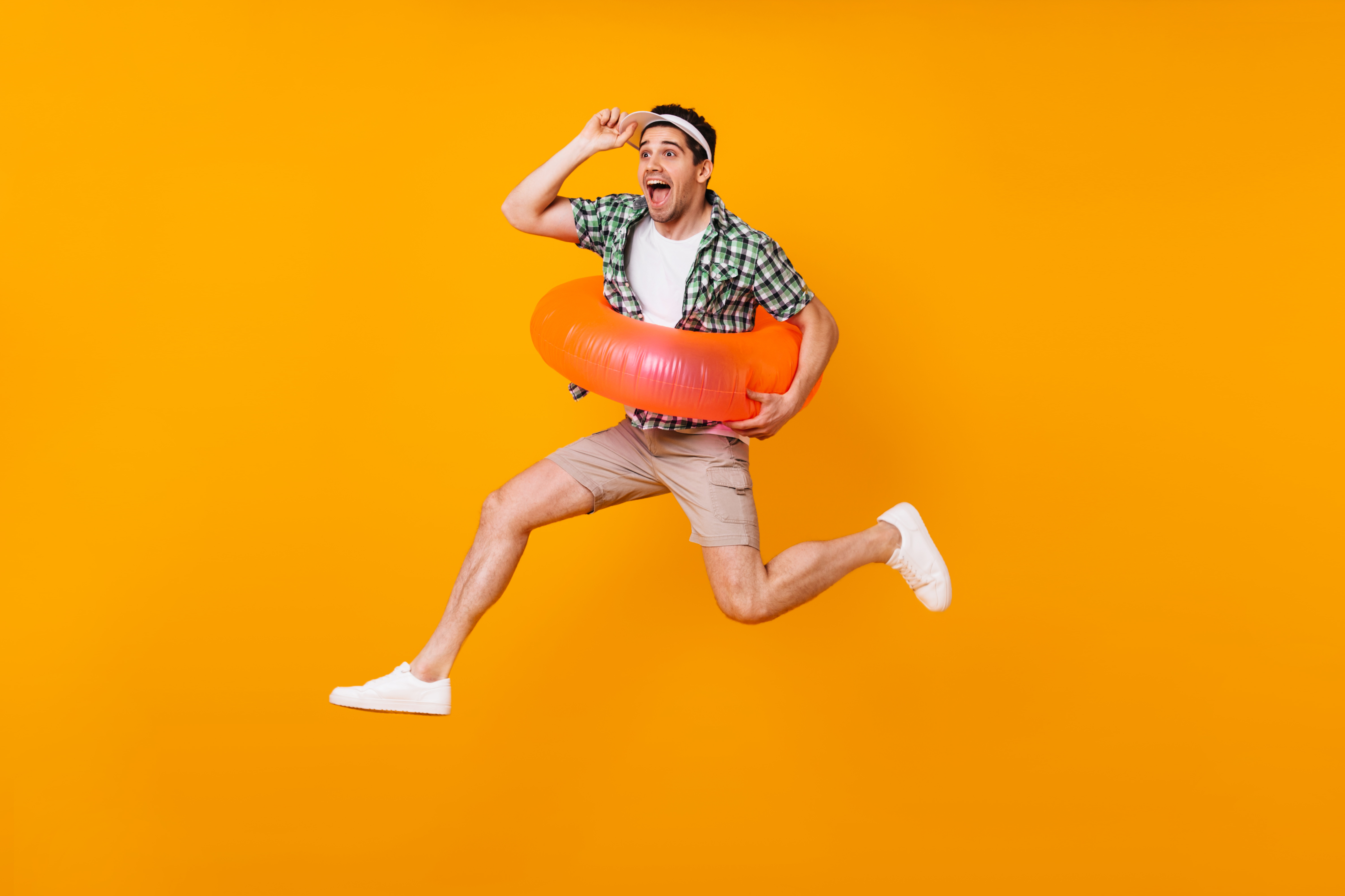 Emotional guy in high spirits jumps on orange background with inflatable circle and takes off his cap.